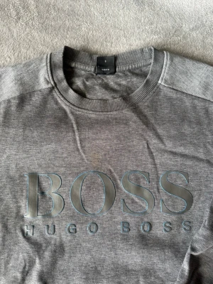 Grå sweatshirt från Hugo Boss - Snygg grå sweatshirt från Hugo Boss med stort BOSS-tryck framtill. Tröjan har rund halsringning, långa ärmar och ribbade muddar vid ärmslut och nederkant. Perfekt för en avslappnad och stilren look.