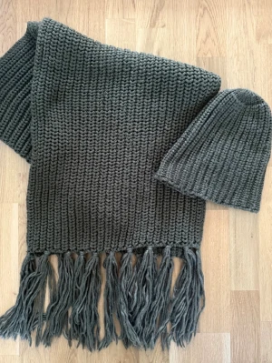 Mörkgrå stickad halsduk med fransar och tillhörande mössa  - En chunky mörkgrå stickad halsduk med långa fransar i ändarna och med stickad mössa. Halsduken är bred och har ett klassiskt ribbstickat mönster som ger en cool och avslappnad look. Perfekt för att hålla sig varm och stilren under kalla dagar.