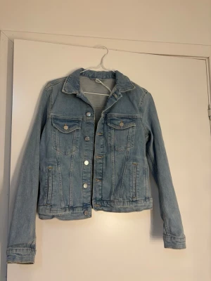 Ljusblå klassisk jeansjacka - Snygg ljusblå jeansjacka med silverfärgade knappar och två bröstfickor med lock. Jackan har en klassisk krage, raka ärmar och markerade sömmar framtill. Perfekt för en avslappnad och trendig look.