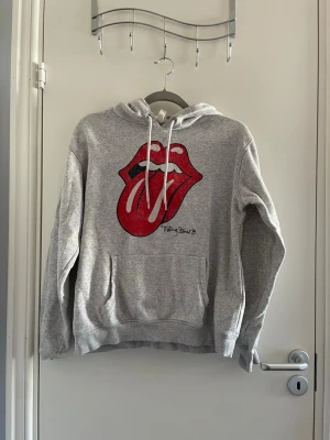 Grå Rolling Stones hoodie med tryck - Grå hoodie med den ikoniska Rolling Stones tungan i rött och vitt på bröstet. Hoodien har dragsko i huvan och en stor magficka framtill. Perfekt för dig som gillar klassiska band och vill ha en bekväm och snygg tröja.