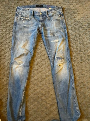 Blå slitna jeans från Replay - Säljer ett par blå jeans från Replay med skinny fit och slitna detaljer. Jeansen har flera hål och slitningar framtill, klassisk femficksmodell och knappgylf. Perfekta för en avslappnad och trendig look.