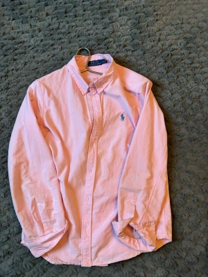 Rosa skjorta från Polo Ralph Lauren - Klassisk rosa skjorta från Polo Ralph Lauren med button-down krage och blå broderad logga på bröstet. Skjortan har lång ärm, knappar framtill och är tillverkad i bomull. Perfekt för en stilren look.