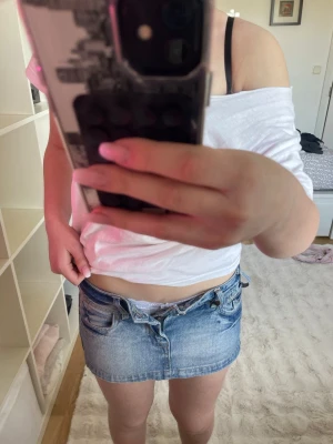 Blå jeanskjol med dubbla knappar - Säljer en kort blå jeanskjol med dubbla knappar och klassiska fickor fram och bak. Kjolen har bälteshällor och en snygg tvättad denimlook. Perfekt för dig som gillar en avslappnad och trendig stil.