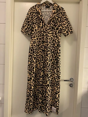 Leopardmönstrad långklänning Asos - Cool långklänning från Asos Design med leopardmönster i beige och svart. Klänningen har korta ärmar, krage och v-ringning. Perfekt för dig som vill sticka ut med en trendig look. Stretchigt material. Oanvänd, prislappen är kvar.