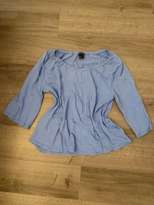 Ljusblå offshoulder-blus från Lindex - Supersöt ljusblå blus från Lindex med offshoulder-modell och trekvartsärmar. Blusen har resår vid halsringningen och är tillverkad i ett mjukt, lätt tyg som ger en avslappnad och luftig känsla.
