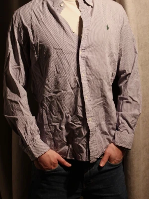 Rutig skjorta från Ralph Lauren - Säljer en klassisk rutig skjorta från Polo Ralph Lauren med diskret lila och vit färg. Skjortan har långärmad design, knappar framtill och ikonisk logga broderad på bröstet. Perfekt för dig som gillar stilrena plagg med snygg passform.