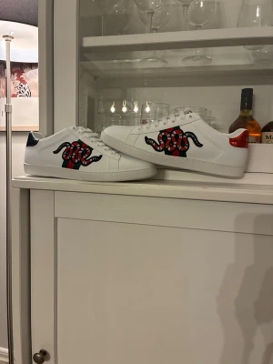 Gucci Ace sneakers med orm - Säljer ett par vita Gucci Ace sneakers i läder med ikonisk broderad röd och svart orm på sidan. Skorna har metallicröd och mörkblå hälflik samt klassisk låg profil. Perfekta för dig som vill sticka ut med lyxig streetstyle. Priset för att diskutera .