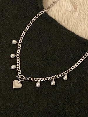 Silverfärgat halsband med pärlor och hjärta - Ett silverfärgat rostfritt halsband med kedja och sex hängande vita pärlor samt ett hjärtformat hänge. Halsbandet har en toggle-låsning och en stilren design som passar perfekt för att ge en söt touch till din outfit.
