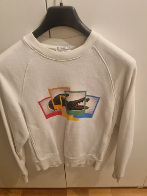 Vit sweatshirt från Lacoste - Vit sweatshirt från Lacoste med klassiskt krokodilmärke på bröstet, inramat av färgglada rutor i rött, gult, grönt och blått. Tröjan har rund halsringning och långa ärmar. 