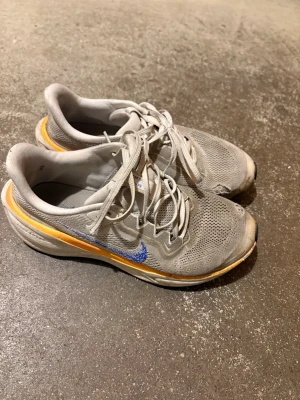 Nike Prototype grå sneakers - Nike Prototype sneakers i grå mesh med blå Swoosh och orange detalj längs sulan. Lågt skaft och ventilerande ovandel. Klassisk snörning och sportig design, perfekt för dig som gillar en clean och modern look.