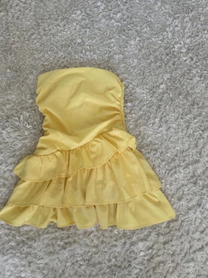 Gul axelbandslös volangklänning - Söt gul axelbandslös kortklänning med flera volanger i kjolen. Klänningen har en figurnära passform upptill och är tillverkad i ett lätt tyg som ger en luftig känsla. Perfekt för varma dagar och somriga tillfällen.