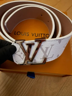 Vitt bälte med metal spänne  - Snyggt vitt bälte med det klassiska monogrammet i ljusgrått och ett stort metallspänne. Bältet har en exklusiv känsla som lyfter vilken outfit som helst.