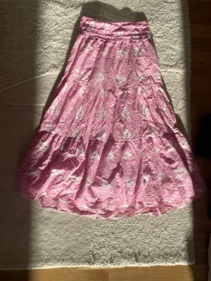 Rosa blommig långkjol med volang - Söt rosa långkjol med blommigt mönster och stretch i midjan. Kjolen har volangdetaljer och är tillverkad i ett lätt, luftigt material med innerfoder. Perfekt för dig som gillar en romantisk och avslappnad stil.