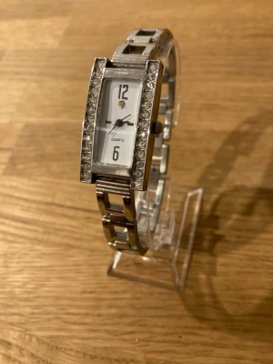 Elegant damklocka med strassdetaljer, Quartz - Elegant damklocka med rektangulär urtavla och strassdetaljer längs sidorna. Urtavlan är vit med siffrorna 12 och 6, samt en hjärtformad markör vid 12. Klockan har ett silverfärgat länkarmband. Baksidan är märkt 'STAINLESS STEEL BACK'. Klockan drivs av ett Quartz-urverk. Fint skick enligt bilder, behöver bara nytt batteri.  Begagnat värde: < 500 SEK Mitt pris: 189 SEK  Skickas alltid snabbt och spårbart. Skriv vid frågor och se gärna mina andra annonser! 