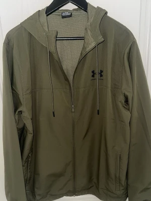 Under Armour jacka  - Snygg olivgrön vindjacka från Under Armour med dragkedja framtill, huva med dragsko och två sidofickor. Jackan har en sportig look och är tillverkad i ett lätt, vindtätt material som passar perfekt för blåsiga dagar.har använt den 1 till 3  gånger brara 