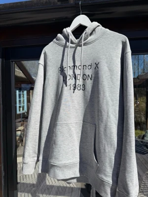 Richmond X hoodie - Snygg grå hoodie från Richmond X med svart text på bröstet. Klassisk modell med huva, dragsnören och stor magficka. Perfekt för en avslappnad streetstyle-look. Få använd, perfekt skick. Storlek S men lite oversized. Skriv vid frågor!