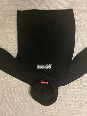 Svart Supreme hoodie med box logo - Svart hoodie från Supreme med klassisk box logo i vitt och svart på bröstet samt röd Supreme-tag på huvan. Hoodien har känguruficka och ribbade muddar. Perfekt för dig som gillar streetwear och vill sticka ut med en ikonisk look.