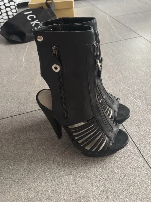 Svarta stövletter med dragkedja - Snygga svarta stövletter med höga klackar och öppna detaljer framtill. Skorna har dragkedjor både fram och på sidan samt coola metallknappar. Tillverkade i konstläder med trendig design som sticker ut.
