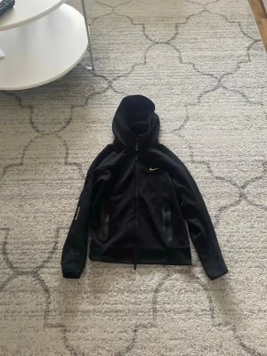 Svart Nike hoodie med dragkedja - Svart hoodie från Nike med hel dragkedja och huva. Swoosh-logga i guld på bröstet och diskreta detaljer på ärmen. Två fickor framtill med dragkedja. Tillverkad i mjukt material som passar perfekt för en sportig och avslappnad stil.