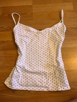 Vit prickig linnetopp Gina Tricot - Supersöt vit linnetopp från Gina Tricot med svarta prickar och tunna axelband. Toppen har en lätt v-ringad halsringning och är tillverkad i mjuk bomull, perfekt för varma dagar eller som layering-piece. Se lagning på de sista två bilderna 💗