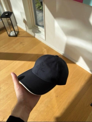 Loro Piana keps - Snygg marinblå keps från Loro Piana med diskret broderad logga framtill. Kepsen har böjd skärm med vit kant och insida i klassiskt rutigt tyg. Tillverkad i Green Storm System-material som står emot väta.