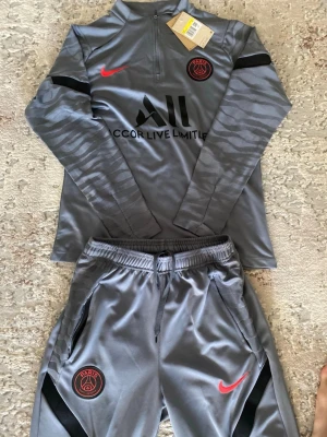 Grå PSG träningsset från Nike - Snyggt grått träningsset från PSG och Nike med långärmad tröja och matchande byxor. Setet har röda Nike-loggor och PSG-emblem, samt svarta detaljer och dragkedja vid halsen. Materialet är mjukt och stretchigt, perfekt för träning eller chill.