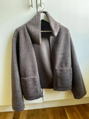 Brun teddyjacka med stora fickor - Mysig brun  teddyjacka med oversized krage och två stora framfickor. Jackan har rak passform och dragkedja framtill. Perfekt för kalla dagar när du vill vara både varm och trendig. Tillverkad i fluffigt teddy-material som ger en cool och avslappnad look.