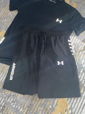 Svart träningsset från Under Armour - Säljer ett svart träningsset från Under Armour med t-shirt och shorts. Båda plaggen har logotypen tryckt i vitt och texten 'ARMOUR' på sidan. Materialet är lätt och andas, perfekt för gym eller löpning.