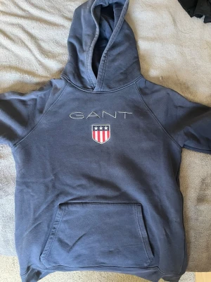 Blå hoodie från GANT med logga - Snygg blå hoodie från GANT med broderad logga och amerikanskinspirerat sköldmärke på bröstet. Klassisk känguruficka framtill och stor huva. Tillverkad i mjukt bomullsmaterial som är perfekt för chill dagar. Enkel och stilren design som funkar till det mesta.