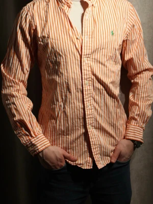 Randig skjorta från Polo Ralph Lauren - Snygg långärmad skjorta från Polo Ralph Lauren med orange och vita ränder. Klassisk krage, knäppning framtill och den ikoniska gröna loggan broderad på bröstet. Tillverkad i mjuk bomull för en bekväm känsla och stilren look.