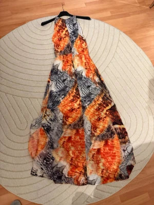 Långklänning med orange mönster - Snygg långklänning med ett unikt mönster i orange, svart, grått och vitt. Klänningen har halterneck och ett svepande fall som ger en elegant siluett. Perfekt för dig som vill sticka ut med färg och mönster. Materialet känns lätt och luftigt.