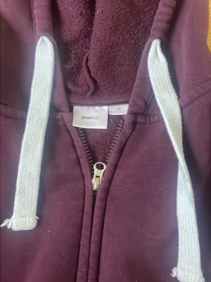 Lila hoodie från Gina Tricot - Mysig vinröd hoodie från Gina Tricot med dragkedja framtill och stora fickor. Tröjan har vita snören i huvan och ribbade muddar vid ärmslut och midja. Perfekt för en avslappnad stil och enkel att matcha med jeans eller leggings.