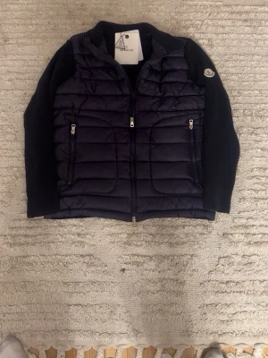 Mörkblå cardiganjacka från Moncler - Snygg mörkblå cardiganjacka från Moncler med vadderad framsida och stickade ärmar och rygg. Jackan har två dragkedjeförsedda fickor fram och en Moncler-logga på ärmen. Perfekt för lager-på-lager och en stilren look.