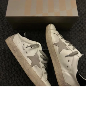 Golden Goose Superstar vita sneakers - Golden Goose Superstar sneakers i vitt läder!!! grå stjärndetalj på sidan, grå skosnören och svart hälflik. Klassisk låg modell med slitna detaljer. Funkar bra nästan överallt. KOM GÄRNA MED PRISFÖRSLAG, ÖPPEN FÖR ALLT🤩🤩