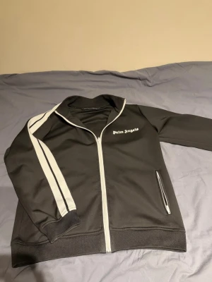  Palm Angels kofta  - Säljer en svart zip kofta från Palm Angels med vita detaljer och två vita ränder längs ena ärmen. Hoodien har dragkedja framtill, ribbade muddar och fickor med dragkedja. Materialet är mjukt och stretchigt, perfekt för en avslappnad streetwear-look. Kan passa för M 