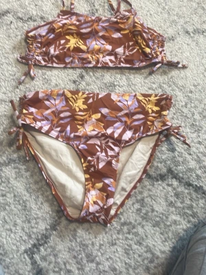 Brun blommig bikini med snörning - Snygg bikini med blommigt mönster i brunt, gult och lila. Överdelen har smala axelband och snörning i sidorna, och underdelen är högt skuren med knyt i sidorna. Perfekt för strandhäng och sommarens bad.