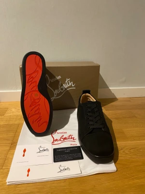 Christian Louboutin skor  - Snygga svarta sneakers från Christian Louboutin med klassisk röd sula och diskret logga på hälen. Skorna har en låg profil, svarta skosnören och är tillverkade i mocka och läder. Perfekta för dig som vill ha en exklusiv och stilren look.