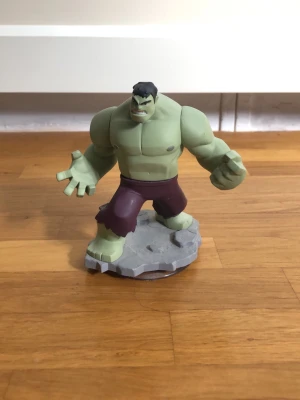 Intressekoll! Hulk figur på grå bas - Cool Hulk figur i grönt och lila, står på en grå platta. Figuren är gjord av plast och har muskulös kropp med trasiga lila shorts. Perfekt som samlarobjekt eller dekoration för Marvel-fans.