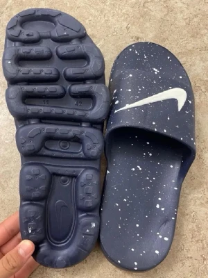 Blå Nike sandaler med vit splash - Mörkblå sandaler från Nike med vit splash-effekt och stor vit swoosh-logga på ovandelen. Tillverkade i ett lätt och bekvämt syntetmaterial med unik, mönstrad sula för extra grepp. Perfekta för chill dagar eller efter träningen.