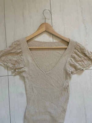 Beige ribbad topp med puffärm - Supersnygg beige topp med ribbad kropp och v-ringning. Ärmarna är korta, puffiga och har ett genombrutet mönster som ger en unik look. Perfekt till jeans eller kjol för en trendig outfit.