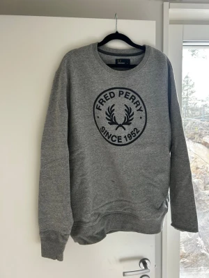 Grå sweatshirt från Fred Perry - Klassisk grå sweatshirt från Fred Perry med stort svart tryck på bröstet och texten 'Fred Perry Since 1952'. Tröjan har rund halsringning, ribbade muddar och är tillverkad i mjukt bomullsmaterial. Perfekt för en avslappnad och stilren look.
