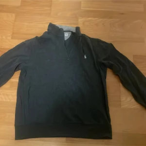 Mörkgrå half zip tröja från Polo Ralph Lauren - Mörkgrå långärmad tröja från Polo Ralph Lauren med klassisk half zip och hög krage. Tröjan har en broderad logga på bröstet och är tillverkad i mjuk bomull. Perfekt för lager-på-lager och chill stil.