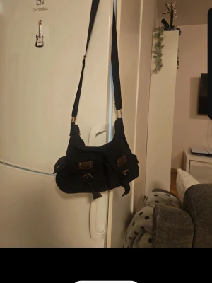 Svart axelväska med fickor - Snygg svart axelväska i tyg med två framfickor som har spännen och detaljer i brunt läderimitation. Väskan har justerbar axelrem och en mjuk, lite rundad form. Perfekt för dig som gillar praktiska detaljer och stilren design.