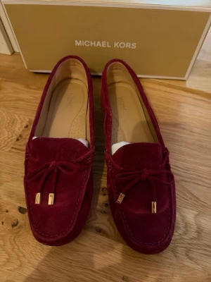 Vinröda loafers från Michael Kors - Snygga vinröda loafers i mocka från Michael Kors med dekorativa rosetter och guldfärgade detaljer framtill. Klassisk seglarsko-modell med låg klack och mjuk innersula för extra komfort. Perfekta för dig som vill ha en stilren och trendig look.