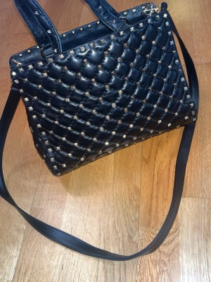 Svart Valentino väska (nyskick) - Snygg svart handväska i quiltad design med små silvriga nitar över hela väskan. Väskan har både handtag och en lång axelrem så du kan bära den på olika sätt. Tillverkad i konstläder med fyrkantig form och coola detaljer. Märket från valentino, 100% äkta och ny skick.