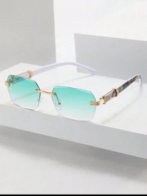 Grönblå kantlösa solglasögon - Snygga kantlösa solglasögon med fyrkantiga glas i grönblå toning. Bågarna har gulddetaljer och transparenta skalmar med marmorerad effekt. Perfekta för dig som vill sticka ut med en modern och trendig look.
