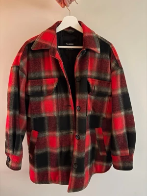 Röd och svart rutig overshirt Pull&Bear - Overshirt i rött och svart rutigt mönster från Pull&Bear. Klassisk skjortkrage, stora knappar framtill och två bröstfickor med lock. Tillverkad i ett tjockare, mjukt material som ger en avslappnad och trendig look.