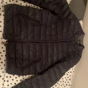 Svart pufferjacka från Calvin Klein - Säljer en svart pufferjacka från Calvin Klein med quiltad design och två snedställda dragkedjefickor framtill. Jackan har hög krage och diskret logotyp på bröstet. Perfekt för kalla vinterdagar, tillverkad i syntetmaterial.