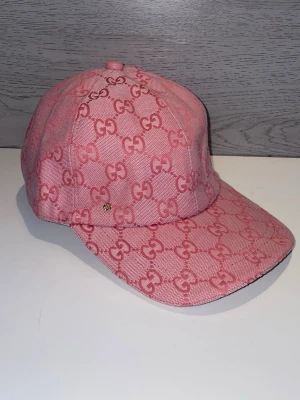 Rosa Gucci keps med GG-mönster - Säljer en rosa keps från Gucci med ikoniskt GG-mönster över hela kepsen. Kepsen har böjd skärm och är tillverkad i ett slitstarkt tygmaterial. Perfekt för dig som vill ha en trendig accessoar med lyxig känsla och tydlig logga.