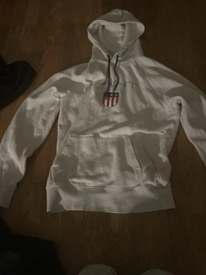 Vit hoodie från Gant med logga - Vit hoodie från Gant med broderad logga och ett emblem med stjärnor och ränder på bröstet. Hoodien har en stor huva med snören och en klassisk magficka. Materialet är mjukt och bekvämt, perfekt för en avslappnad stil.